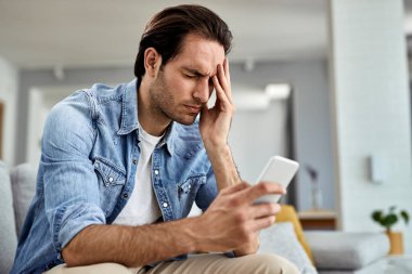 Young man using smart phone and holding his head in pain at home.