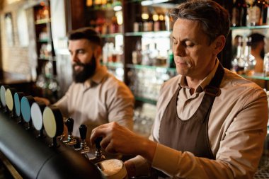 Mid adult bartender and his coworker working in a pub and pouring beer from a beer tap.