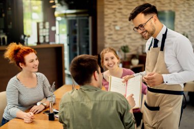 Happy waiter showing menu to a guest who came with friends in a cafe. 