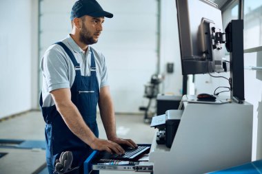 Araba atölyesinde çalışırken bilgisayar kullanan oto tamircisi. 