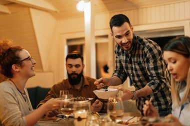 Small group of happy friends having dun during dinner at home. Focus is on man serving food. 