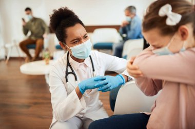 Little girl with face mask sneezing into elbow while talking to African American female dentist in waiting room. Focus is on dentist. 