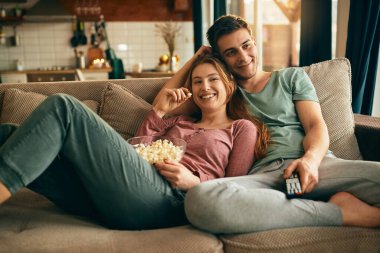 Young happy couple watching movie on TV and eating popcorn while relaxing in the living room. 