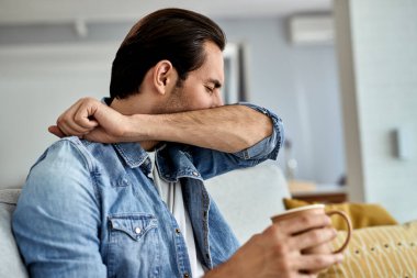 Young man sneezing into elbow while drinking teat in the living room. 