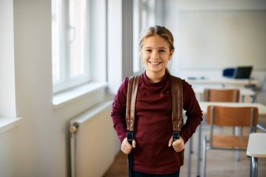 Happy little girl with backpack standing in classroom at elementary school and looking at camera. 