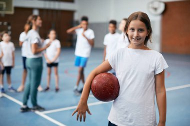 Happy elementary student holding volleyball ball while having PE class at school gym Her friends and coach are in the background.