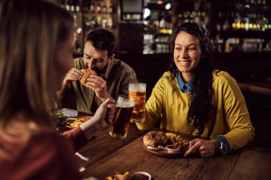 Young happy woman and her friend toasting with beer while eating burgers and French fries in a pub. 