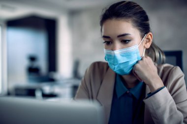 Young businesswoman working on laptop while wearing face mask in the office due to coronavirus pandemic.