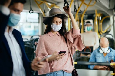 Young woman using smart phone and wearing face mask while traveling by public transport during coronavirus pandemic.