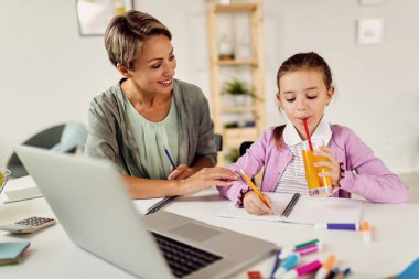 Small girl drinking orange juice while writing in notebook and learning at home with her mother. 