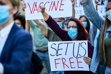 Crowd of displeased people wearing protective face masks while protesting on the streets during coronavirus pandemic. Focus is on woman with raised fist carrying a placard with set us free inscription. 