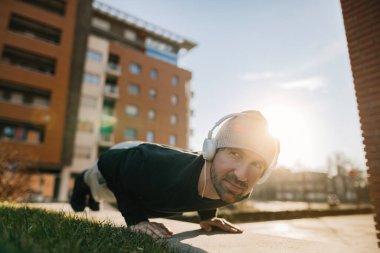 Smiling sportsman exercising on the street and doing push-ups while listening music over headphones.
