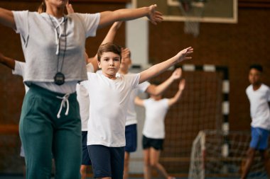Group of school children and sports teacher doing warm-up exercises during PE class at elementary school gym. Focus is on boy with arms outstretched. 