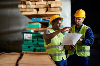 African American worker and his colleague discussing while analyzing paperwork at wood warehouse.