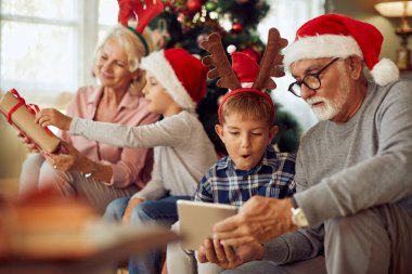 Senior man and his grandson using touchpad while his wife and other grandson are opening Christmas presents in the background at home.