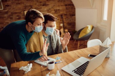 Couple greeting someone during video call while wearing face masks at home due to coronavirus pandemic. 