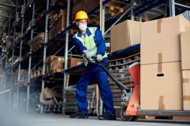 Warehouse worker wearing protective face mask and pulling packages on pallet jack at storage compartment. 