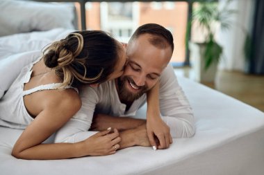 Affectionate woman kissing her boyfriend while relaxing on bed in the morning. 