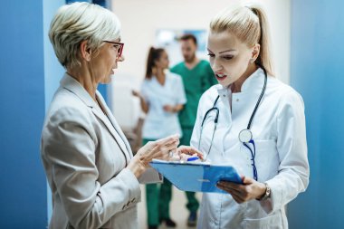 Senior woman communicating with a doctor who is analyzing her medical reports in a hallway at the hospital. 