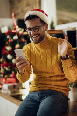 Young happy man waving while making video call over smart phone on Christmas Eve at home. 