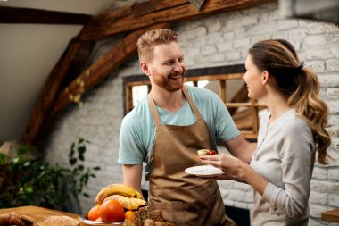 Happy man talking to his girlfriend who is eating small sandwich for breakfast in the kitchen. 