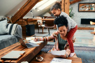 Busy African American mother working at home while her playful daughter is climbing on her shoulder and distracting her from work.