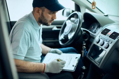 Aracın içini inceleyen ve bir atölyedeki panoya veri yazan oto tamircisi.