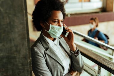 Korumacı yüz maskeli Afrikalı bir iş kadını, tren istasyonunda halka açık bir salondayken akıllı telefondan iletişim kuruyor.. 