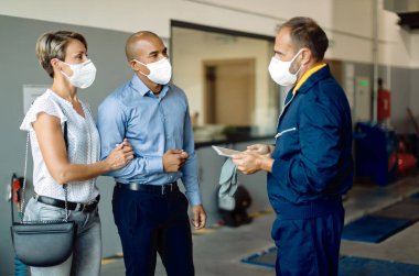 African American man and his wife communicating with their car mechanic in a workshop. They are wearing face masks due to coronavirus pandemic. 