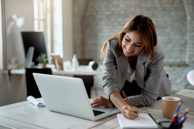 Happy businesswoman writing notes while using laptop and working in the office. 