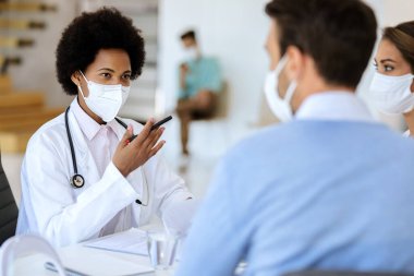 African American doctor having an appointment with a couple and wearing protective face mask while communicating with them at doctor's office. 