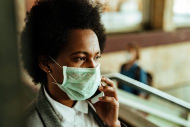 Thoughtful black businesswoman wearing protective mask while communicating on cell phone during virus epidemic at airport corridor. 