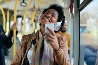 Happy African American woman using smart phone and enjoying in music over headphones while commuting by public transport. 