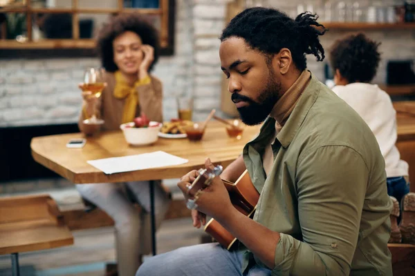 Black man playing acoustic guitar while his wife and son are sitting in the background at home.
