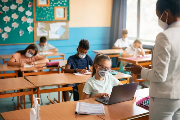 Group of students and their teacher wearing protective face masks at elementary school. Focus is on girl using laptop during a class.