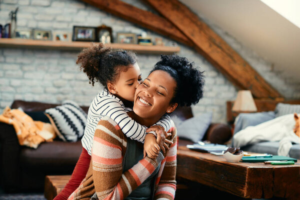 Happy African American mother enjoying while daughter is embracing and kissing her at home.