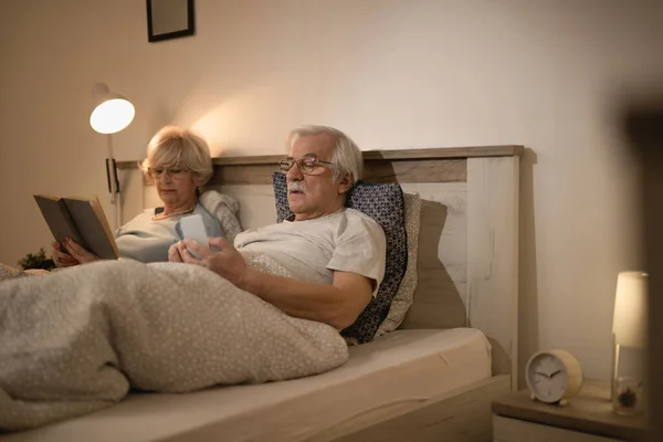 Mature couple relaxing in bed in the evening. Focus is on man using smart phone while his wife is reading a book.