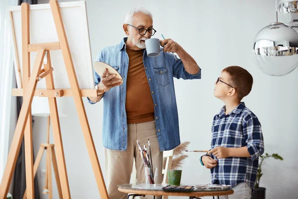 Senior man drinking coffee and talking to his grandson while teaching him to paint on canvas at home.
