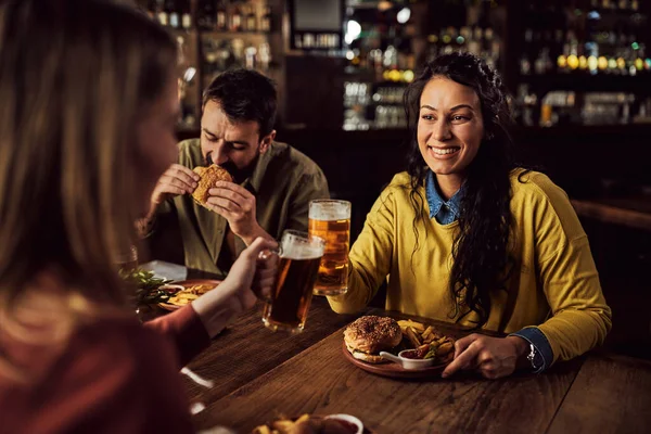Young happy woman and her friend toasting with beer while eating burgers and French fries in a pub.