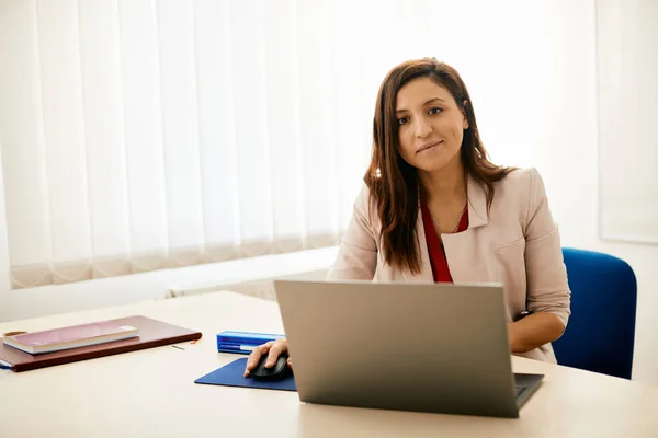 Smiling female professor using computer during a class at school and looking at camera.