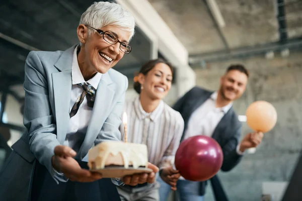 Happy mature businesswoman holding Birthday cake while throwing surprise party with her associates in the office.