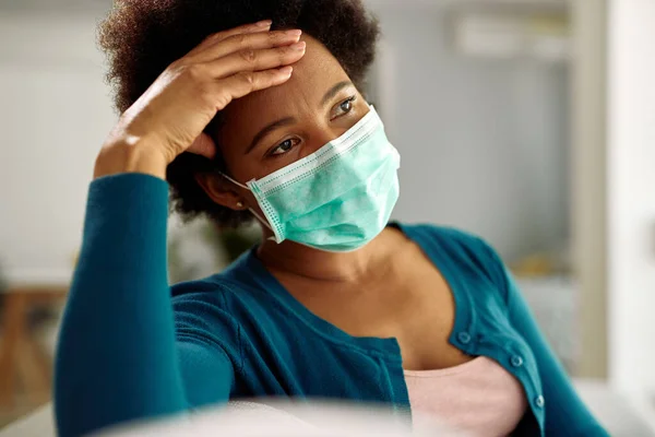 Distraught black woman wearing face mask while sitting at home during virus epidemic.