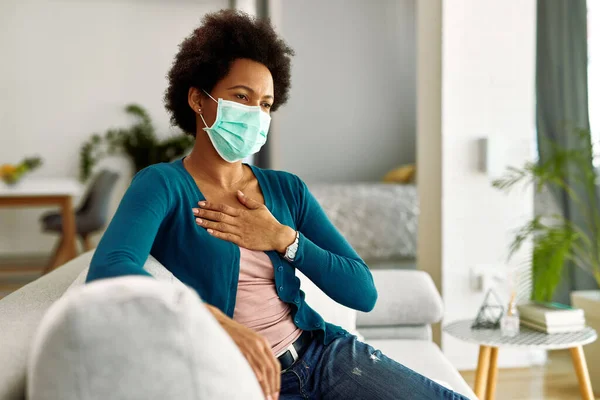 Black woman wearing face mask and holding her chest in pain while sitting on the sofa in the living room.