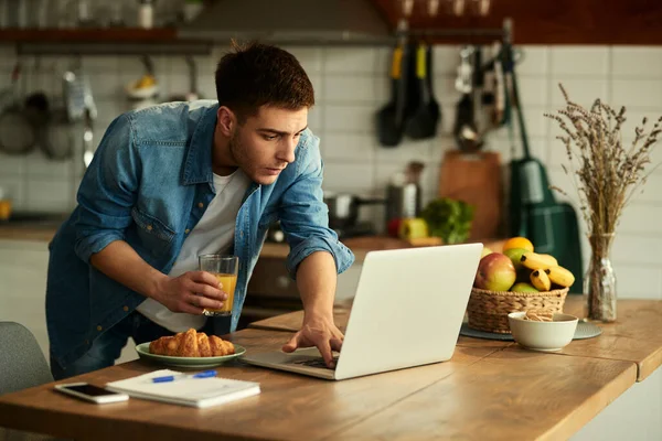 Young man reading an e-mail on laptop while drinking orange juice and working at home.