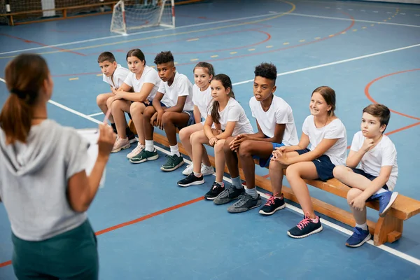 Multi-ethnic group of school kids listening to their sports teacher during physical education class at school gym.