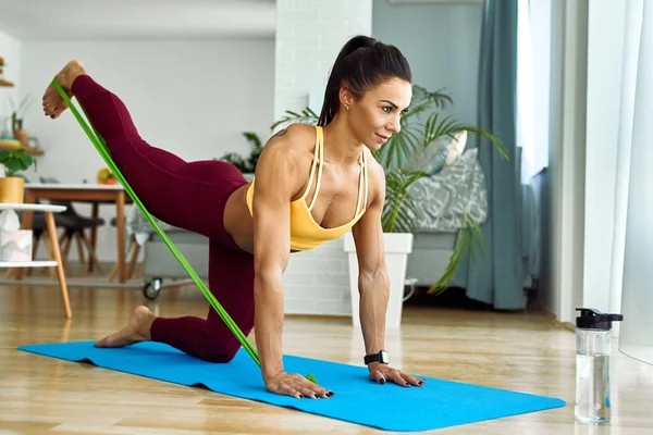 Young sportswoman using a power band while doing leg exercises at home.