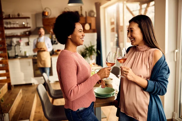 Happy Asian and African American women toasting with wine glasses before the meal at home.
