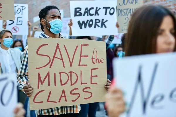 Multi-ethnic crowd of people protesting against unemployment due to coronavirus pandemic. Focus is on black man holding banner with \'save the middle class\' inscription.