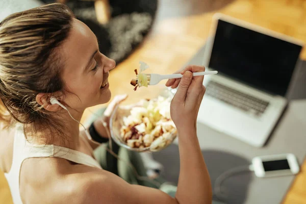 High angle view of happy female athlete eating salad while resting after sports training at home.