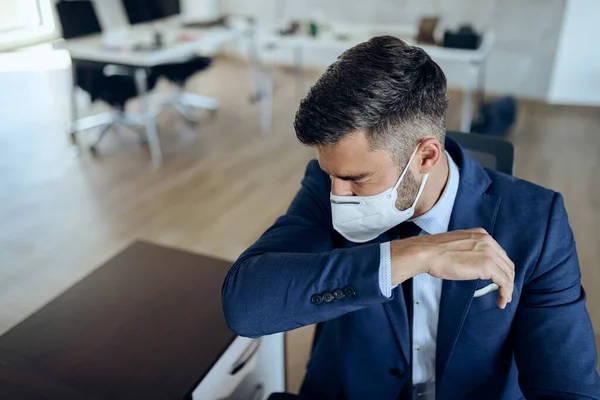Male entrepreneur sneezing into elbow while wearing protective face mask in the office.